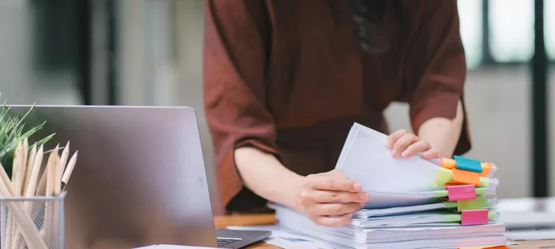 Một nhân viên đang chuẩn bị các giấy tờ với nhiều màu sắc được phân loại và một chiếc laptop trên bàn làm việc
