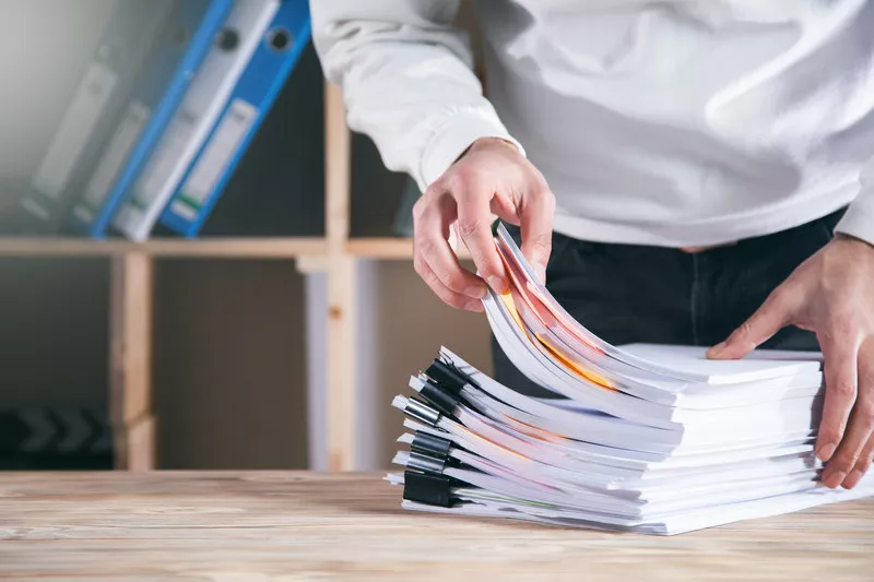 Một người đàn ông đang xem xét và sắp xếp một chồng tài liệu giấy tờ trên bàn gỗ, phía sau là kệ tài liệu