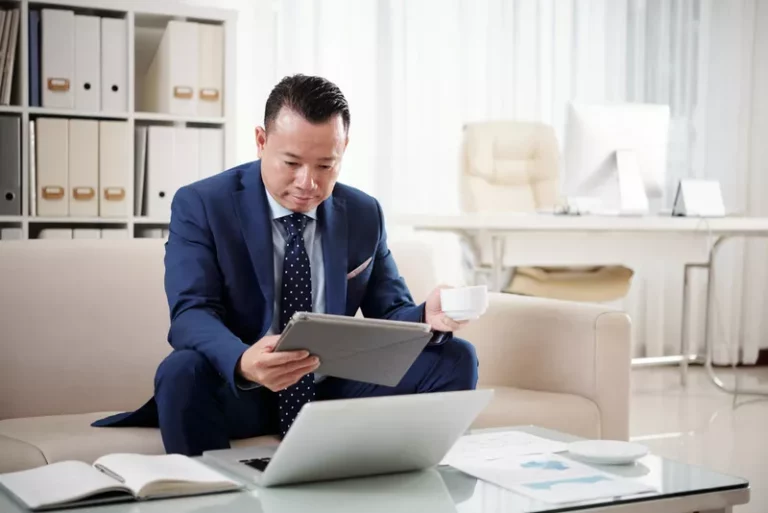 Một người đàn ông doanh nhân châu Á, mặc vest xanh, đang ngồi trên ghế sofa màu kem trong văn phòng